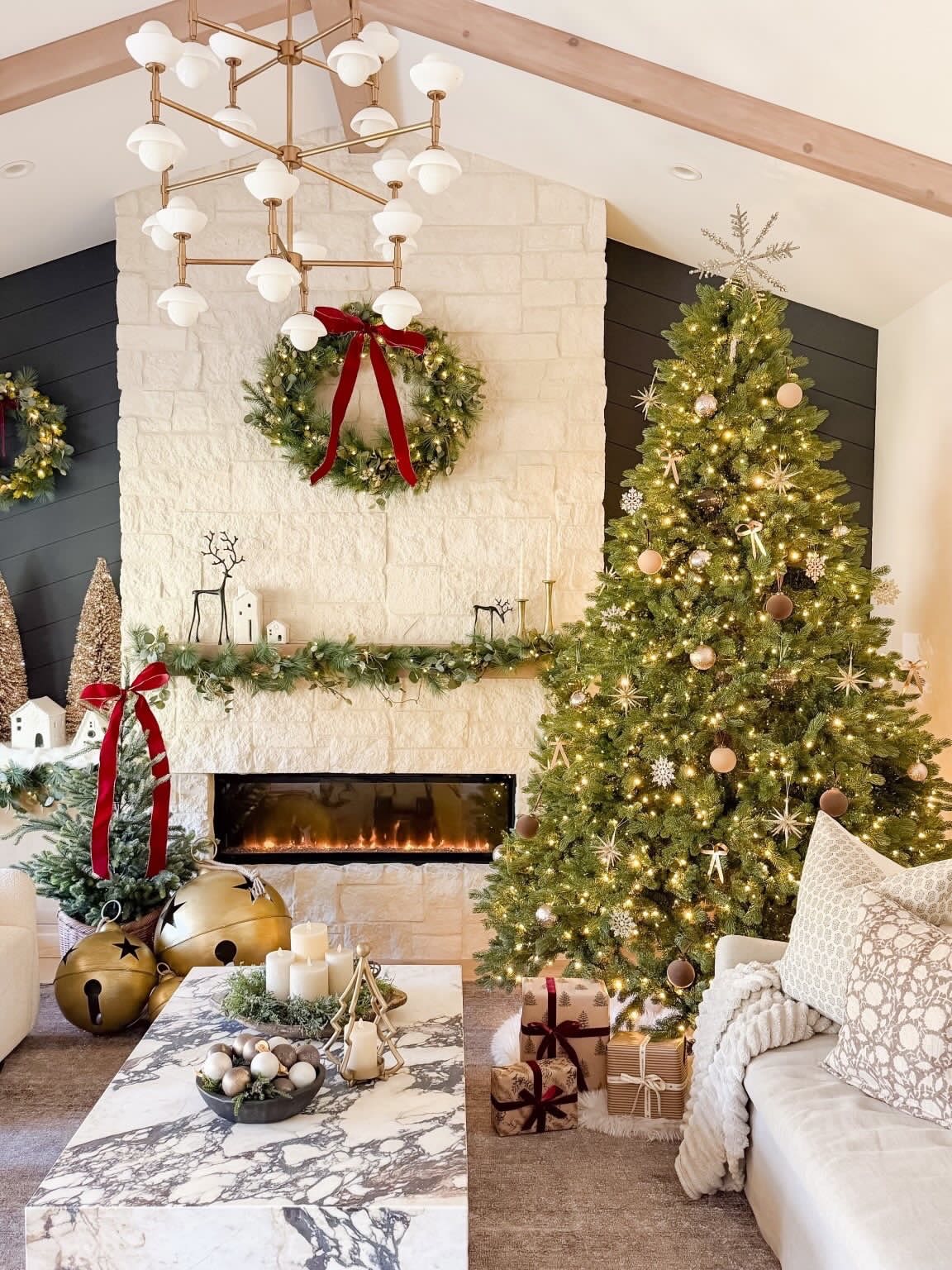 A tall decorated green Christmas tree beside a white brick fireplace with a wreath in a room with a vaulted ceiling.