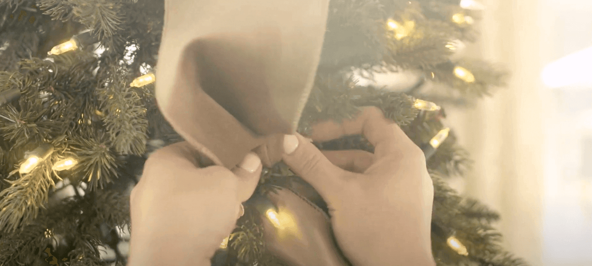 Hands pinching a loop of neutral ribbon into the branches of a lit green Christmas tree.
