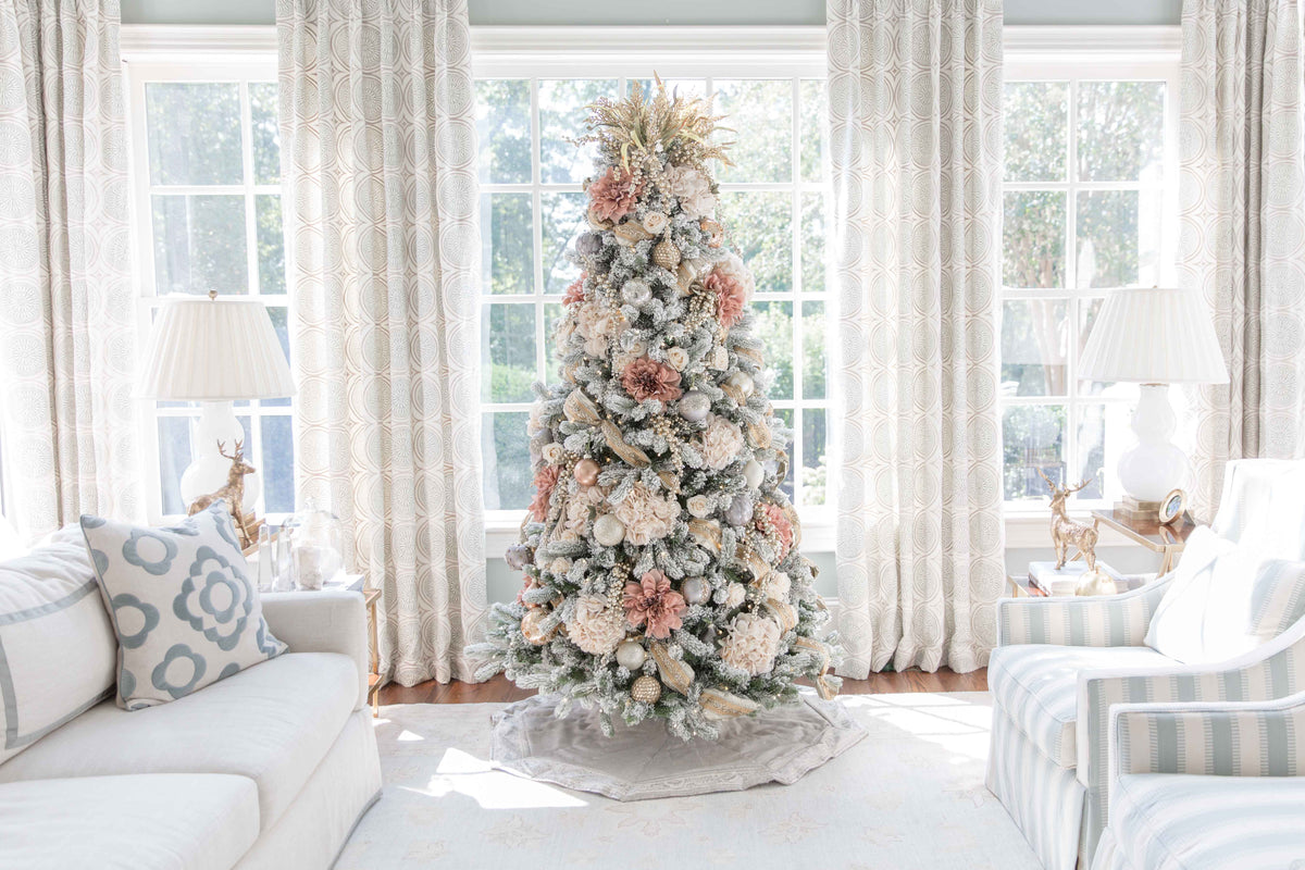 A Christmas tree decorated with pink and cream floral picks and ribbon in a bright living room with white sofas.