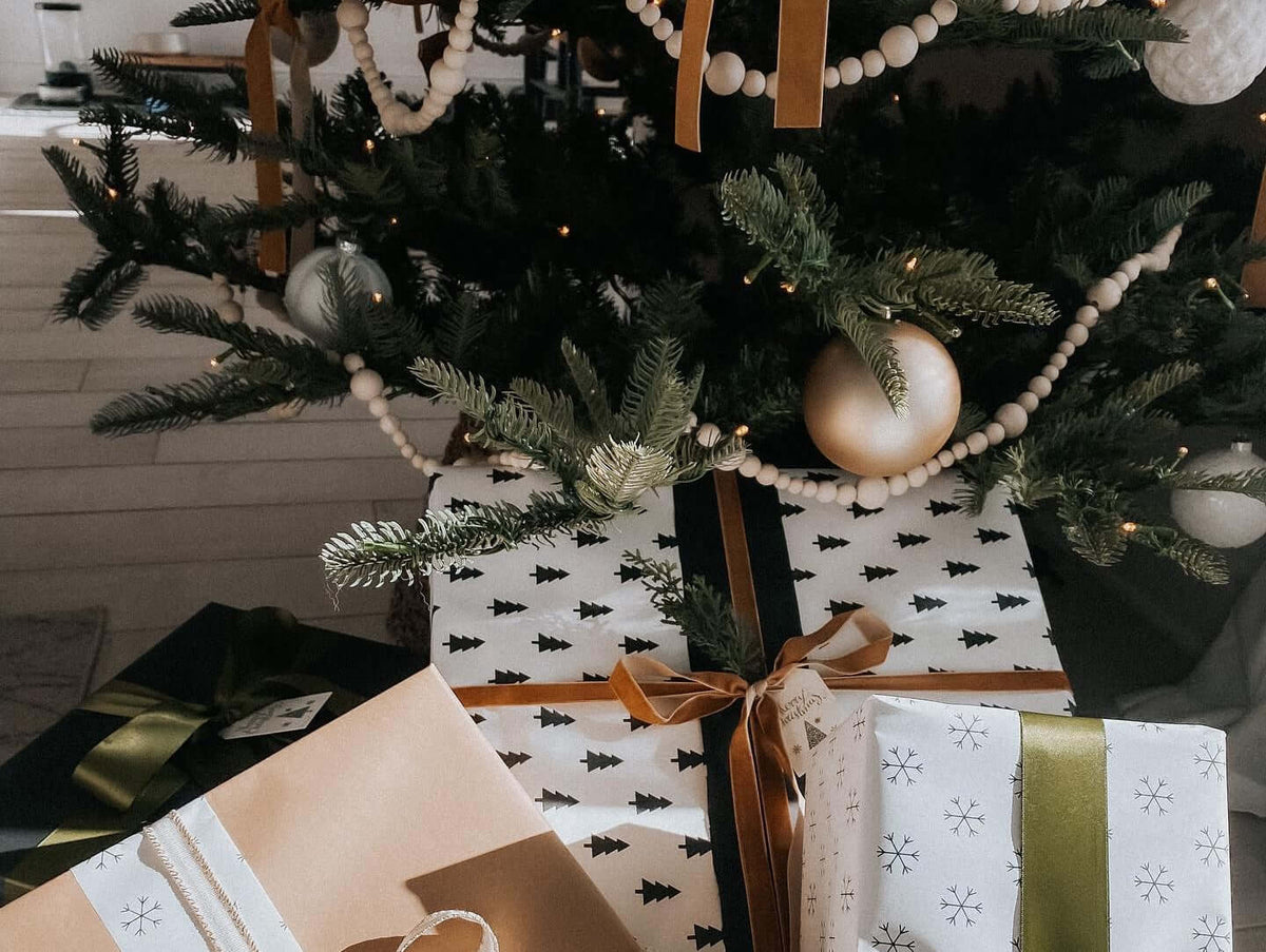 Gifts in tree-print and kraft paper under a green tree decorated with wood bead garland and neutral ornaments.