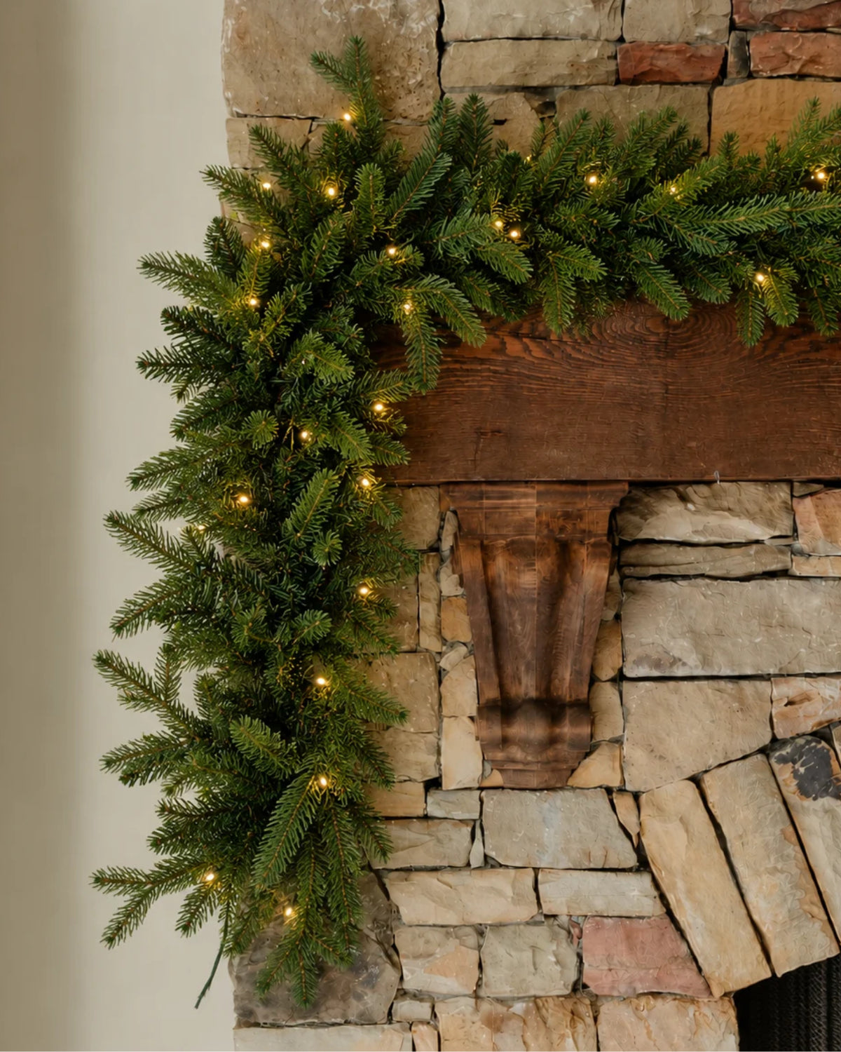 King of Christmas 9' x 16" Alpine Fir Artificial Christmas Garland with warm white LED lights draped elegantly over a rustic wooden mantel and stone fireplace.