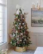 Alpine Fir Slim artificial Christmas tree, pre-lit with warm white LED lights, decorated with a white bow topper and a gold tree collar beside a window